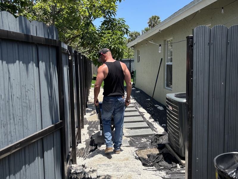 Wheelbarrowing gravel down a narrow side yard between a fence and the house